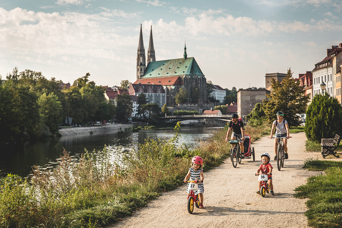 Eine Familie fährt am Neißeufer Rad; im Hintergrund prägen die Türme der Görlitzer Peterskirche und die Altstadt das Stadtbild.
