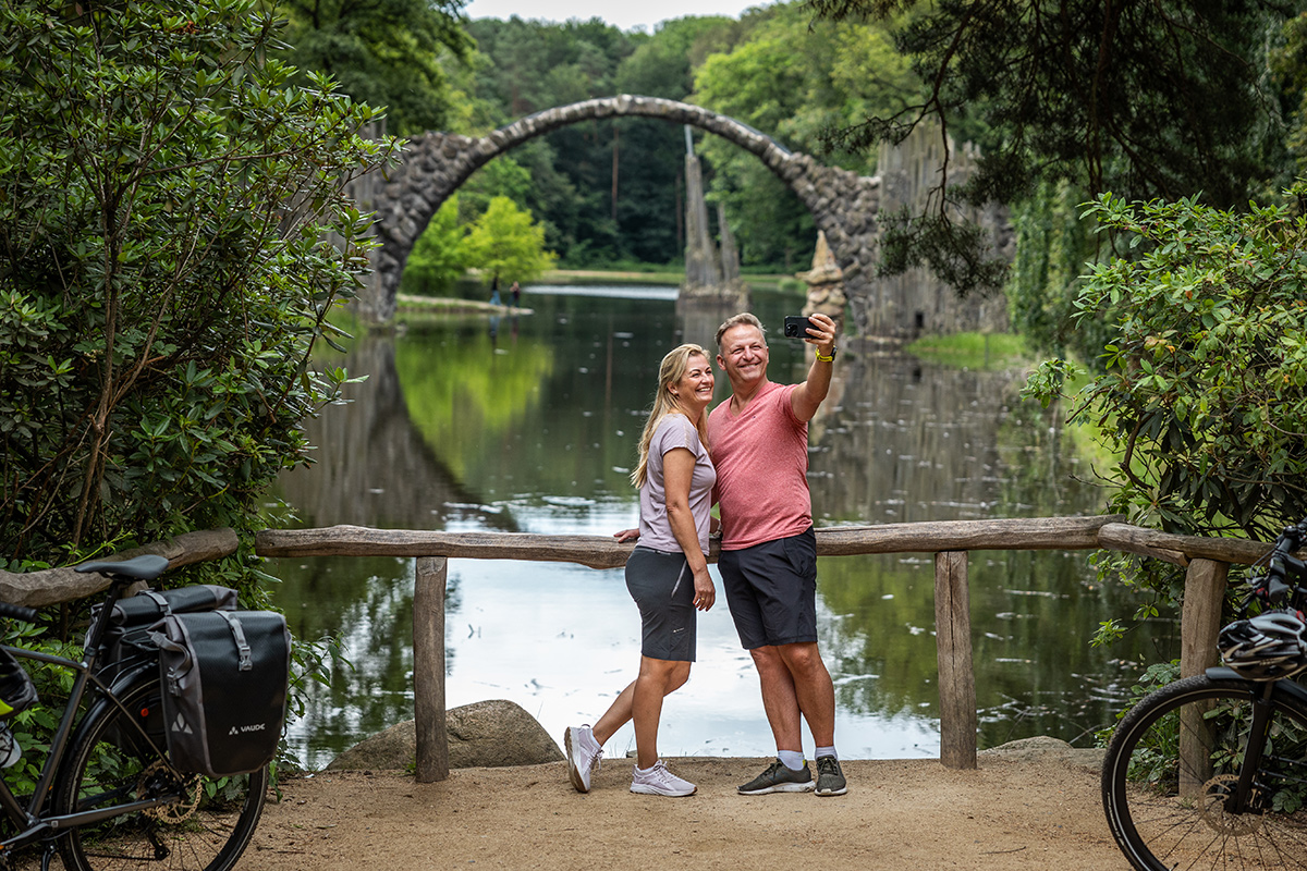 Zwei Radreisende machen ein Erinnerungsfoto vor der Rakotzbrücke im grünen Kromlauer Park.
